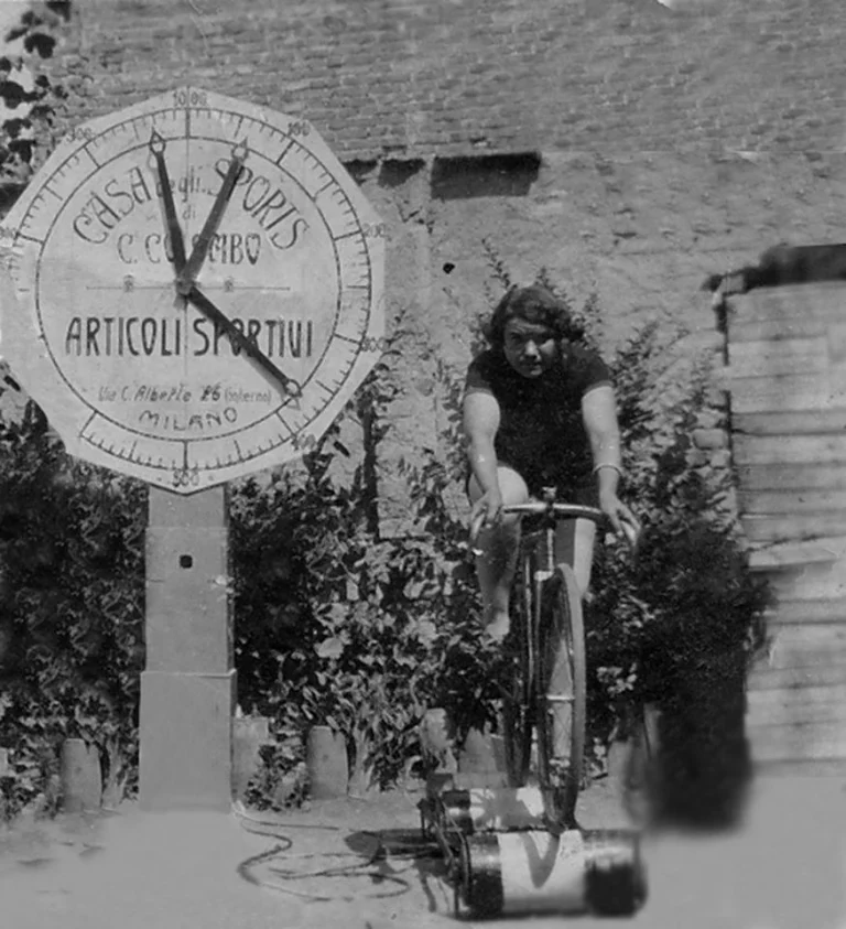 Black and white photo: a young woman in a dark jersey pedals a bicycle set on rollers in front of a brick wall; beside her stands a large dial with hands and the words “Articoli sportivi, Milano”.