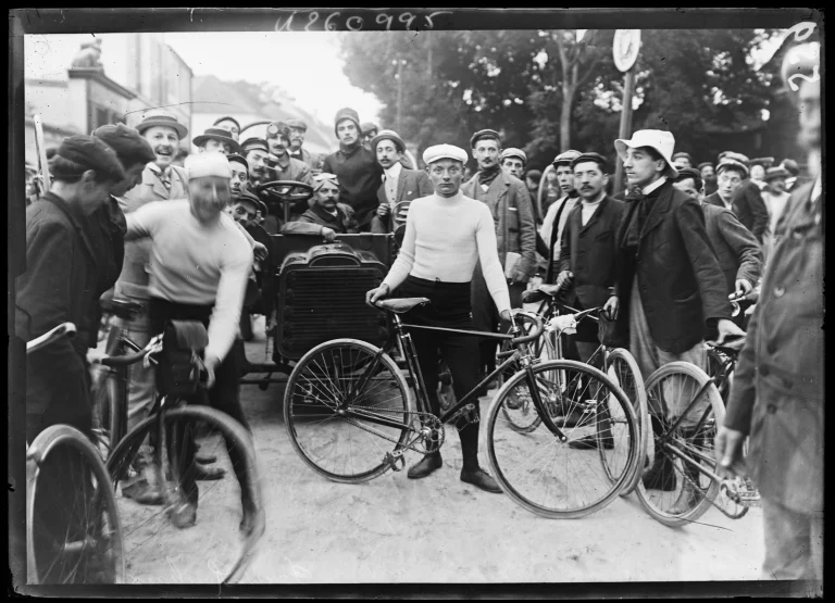 Black and white photo: in the centre a young rider in a white cap and white sweater holds his bicycle, surrounded by a crowd of men in hats and caps, next to an early motor car.