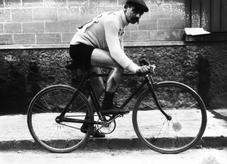 Foto in bianco e nero: un corridore con folti baffi, berretto e maglione chiaro con il numero sulla schiena, seduto in sella a una bicicletta da corsa ferma lungo un marciapiede, davanti a un muro di pietra.